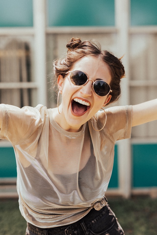 A laughing woman in sunglasses throwing her arms wide on a summer day